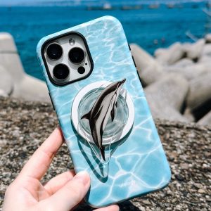 Hand holding the turquoise dolphin case in front of a blue harbour and rocks